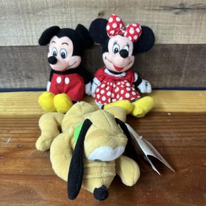 May include: Three Disney plush toys: Mickey Mouse in red shorts, Minnie Mouse in a red polka dot dress, and Pluto the dog. The toys are made of soft fabric and are sitting on a wooden surface.
