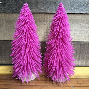 Op de afbeelding: Twee kunstmatige flessenborstelbomen in een levendige magentarode kleur. Elke boom heeft een dicht, getextureerd naaldontwerp en is gemonteerd op een kleine, ronde, lichtgekleurde houten voet, rechtop naast elkaar geplaatst.