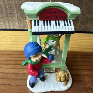 May include: A green and white Christmas bauble featuring a child wearing a blue hat and red jumper standing in front of a music shop called "Tammy's Tune Shop". The shop has a piano keyboard above the door and a snowman inside. The bauble is dated 2015.