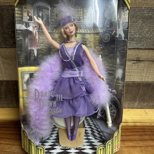 Könnte beinhalten: Collector's Edition Barbie-Puppe in einem lila Flapper-Kleid mit Federboa. Die Puppe befindet sich in einer transparenten Kunststoffbox mit einem schwarz-weißen Schachbrettmuster und einem Vintage-Hintergrund. Die Box hat goldene Akzente.