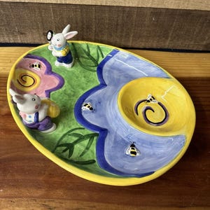 Può includere: Un piatto da portata ovale in ceramica con bordo giallo, con un motivo floreale e a foglie in rosa, verde e blu. Decorato con due statuette di coniglietti e decorazioni a forma di ape. Il piatto ha una piccola ciotola gialla.