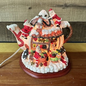 Puede incluir: Una tetera de cerámica con forma de cabaña navideña con figuras de Papá Noel y un techo nevado. La tetera está iluminada con luces blancas cálidas y tiene una base roja.