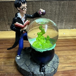May include: A Harry Potter-themed snow globe featuring a figurine of Harry Potter with a book and potion, next to a cauldron with green liquid inside a glass globe. The base is a stone-like texture.