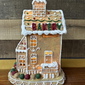 May include: A gingerbread house-shaped decoration with the word "BAKERY" on the roof. The house has white frosting accents, windows, and a chimney. The base features colorful decorative balls. The house is lit from within, creating a warm glow.