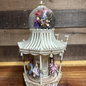 May include: A white and gold carousel snow globe with a clear glass dome. Inside the dome are figurines of people and red confetti. The carousel base features painted horses and riders. The carousel has a decorative roof with small flags.