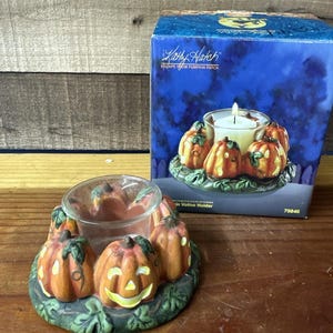 Peut inclure: Un photophore votif en céramique avec des citrouilles oranges et des feuilles vertes. Le support a un insert en verre transparent pour la bougie et est entouré de citrouilles sculptées. La boîte indique "Escape from Pumpkin Patch."