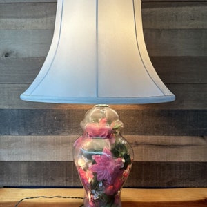 May include: A white lampshade with a clear glass base filled with pink and white flowers. The lamp is on a wooden surface.