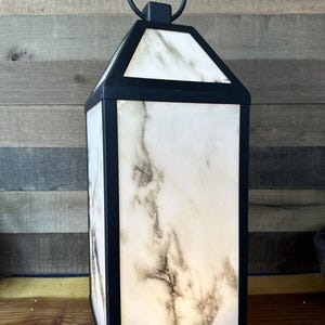 May include: A black metal lantern with a white marble design. The lantern has a handle on top and a clear glass panel on the front.