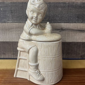 May include: A ceramic figurine of a boy sitting on a barrel. The boy is wearing a shirt and shorts and has a playful expression on his face. The barrel is decorated with a wood grain pattern.