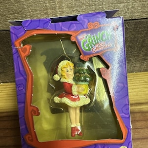 May include: A Cindy Lou Who Christmas ornament in a purple box featuring the text Dr. Seuss How The Grinch Stole Christmas. The ornament depicts a character in a red coat and hat holding a small green tree. The box includes the Universal logo.