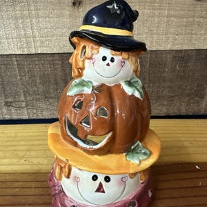 May include: A stacked Halloween-themed ceramic decoration. The top features a witch's hat with a star, a carved pumpkin, and a smiling face. The base is a smiling face with a hat. The colors are orange, navy, yellow, and burgundy.