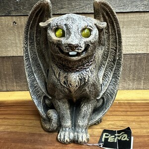 May include: A gray stone-textured winged cat figurine with bright yellow eyes and a chain collar. The cat is in a seated position with wings extended upward. A black tag with the white text "PENO" is attached to the base of the figurine.