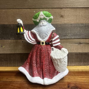 May include: A decorative Santa Claus figurine with a white beard and a green wreath hat. The figure wears a red robe with white trim, a black belt, and carries a lantern and a white basket. The figurine is a Christmas decoration.