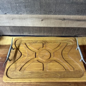 May include: A rectangular wooden serving tray with a carved central design and metal handles. The tray is light brown with a carved sunburst-like pattern in the center. The metal handles are silver and curved.