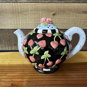 Könnte beinhalten: Dekorative Keramik-Teekanne mit schwarzem Boden und Körper, verziert mit rosa Kirschen und grünen Blättern. Der Deckel ist hellblau mit schwarzen Punkten und einer rosa Rose. Der Griff und der Ausguss sind hellblau mit schwarzen Punkten.