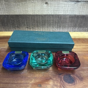 May include: Set of three Marquis Royal Gem votive candle holders in blue, green, and red. Each square-shaped holder has a clear interior and a colored exterior. The set comes in a dark green box.