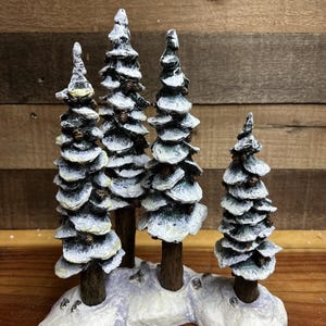 Könnte beinhalten: Eine dekorative Winterlandschaft mit vier schneebedeckten Kiefern. Die Bäume haben braune Stämme und sind mit weißen und grünen strukturierten Ästen geschichtet, die auf einer schneebedeckten Basis ruhen. Der Hintergrund ist eine Holzoberfläche.