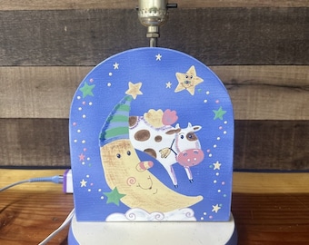May include: A blue and white nightlight shaped like a crescent moon with a cow on top. The moon has a smiling face and the cow has a pink nose. The nightlight is decorated with stars and clouds.