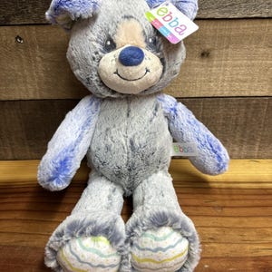 以下が含まれることがあります： グレーとブルーの毛並みのぬいぐるみ犬。犬は青い耳、足、腕をしています。犬のお腹と顔は明るいタン色です。犬には「ebba」と書かれたタグが付いています。犬の足にはカラフルな模様があります。