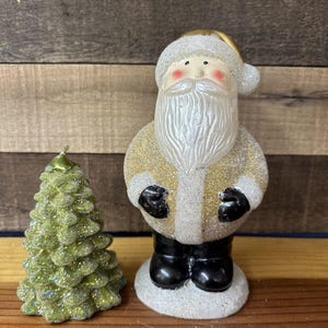 Puede incluir: Un conjunto de dos velas de Navidad, una con forma de Papá Noel y la otra con forma de árbol de Navidad. La vela de Papá Noel lleva un abrigo dorado y blanco con barba blanca y botas negras. La vela del árbol de Navidad es verde con purpurina dorada. Ambas velas están hechas de cera y tienen un acabado brillante.
