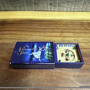 May include: A blue box with "The Nutcracker Suite" text and a ballerina illustration. Inside, a miniature stage with figures is visible. The box is open, revealing a small, detailed diorama of a ballet scene.