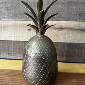 May include: A vintage brass pineapple shaped container with a detailed textured surface. The top of the container is a pineapple top with leaves.