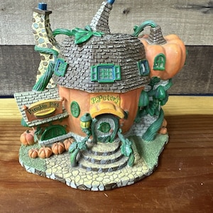 Op de afbeelding: Een grillige, oranje pompoenvormige huisfiguur met een bruin pannendak. Het huis heeft groene accenten, een kleine deur en een bord met de tekst "P. Peter's Pumpkin Pies". Een decoratieve schoorsteen en pompoen maken het ontwerp compleet.