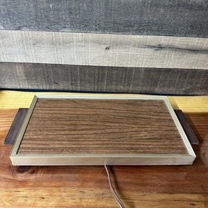 Puede incluir: Una bandeja calentadora eléctrica de madera marrón con un marco de metal plateado y un cable marrón.