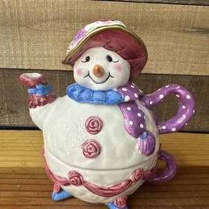 May include: A whimsical snowman teapot with a red and purple hat, scarf, and accents. The white ceramic teapot features a smiling face, rose details, and blue feet. The handle and spout are also purple.