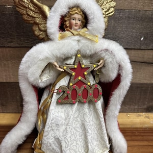 Puede incluir: Figura decorativa de ángel con una capa con capucha ribeteada de piel blanca y alas doradas. El ángel sostiene una estrella roja con el número "2000" en dorado. El vestido del ángel es blanco con un patrón sutil, y la capa tiene un forro rojo.
