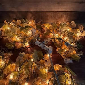 May include: A string of decorative lights featuring artificial autumn-colored maple leaves in shades of yellow, orange, and brown. The strand includes small, clear incandescent bulbs nestled among the foliage, connected by a white electrical cord.