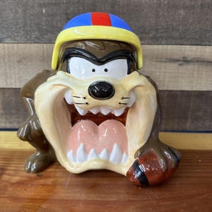 May include: A ceramic figurine of Taz the Tasmanian Devil from the Looney Tunes cartoon. Taz is wearing a blue and yellow helmet with a red stripe and is holding a brown football.