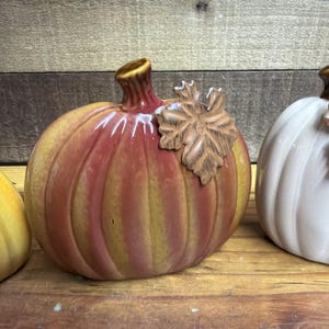 May include: A ceramic pumpkin figurine featuring a glossy finish with vertical stripes in shades of orange, yellow, and brown. The pumpkin has a short, dark stem and a textured, light brown leaf accent attached to the upper side of the body.
