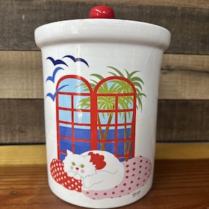 Pode incluir: Jarro de biscoitos de cerâmica branca com tampa vermelha. O jarro apresenta um gato branco com olhos verdes dormindo em um travesseiro de bolinhas rosa e vermelho. O fundo mostra uma janela vermelha com vista para palmeiras e água azul.