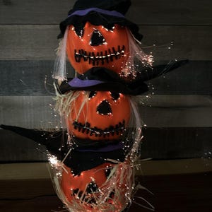 May include: A stacked Halloween decoration featuring three orange pumpkin heads with stitched smiles and black hats. Each pumpkin has a straw-like base and is illuminated with small, twinkling lights, creating a festive, spooky ambiance.