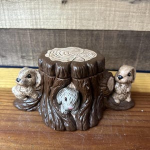 Puede incluir: Caja de baratijas de cerámica marrón con forma de tocón de árbol, con dos cachorros y un conejito. La caja tiene una textura detallada de vetas de madera y una tapa con patrones circulares concéntricos. Los animales son de color marrón claro y gris.