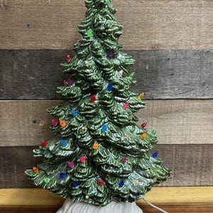 Könnte beinhalten: Ein Weihnachtsbaum aus Keramik mit glänzend grüner Oberfläche. Der Baum ist mit bunten Lichtern geschmückt und steht auf einem weißen Sockel. Ein weißes Netzkabel erstreckt sich von der Basis.