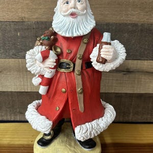 May include: A figurine of Santa Claus wearing a red coat with white fur trim, holding a candy cane and a bottle of Coca-Cola. He is wearing goggles and has a white beard. The figurine is standing on a yellow base with the words "Gary Kraft" written on it.