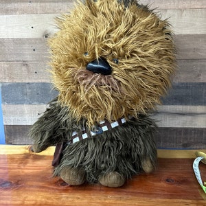 Op de afbeelding: Een Chewbacca pluche pop met bruin en donkergroen haar. De pop heeft een zwarte neus, blauwe ogen en een bruin-witte bandolier. De pop zit op een houten oppervlak.