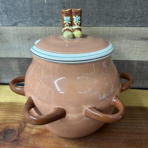 Puede incluir: Una olla de cerámica color melocotón con tapa, adornada con un par de botas de vaquero decorativas. La olla tiene asas marrones y una tapa con rayas turquesas y marrones. Artículo de cocina decorativo.