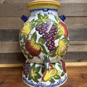 May include: A ceramic drinks dispenser with a lid and tap. The dispenser is adorned with vibrant fruit illustrations, such as apples, pears, and grapes, against a white backdrop. The lid and base feature blue and yellow detailing.