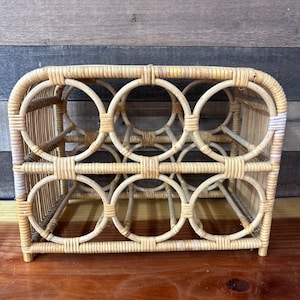 May include: A natural woven rattan wine rack with six circular openings for storing wine bottles. The rack is made of light brown and beige rattan.