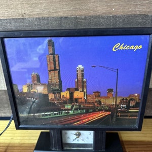 Könnte beinhalten: Schwarze Tischuhr mit einem Chicago-Skyline-Bild und dem Stadtnamen in Gelb. Die Uhr hat ein rechteckiges Zifferblatt mit einer kleinen analogen Uhr an der Basis. Das Bild zeigt die Stadt bei Nacht mit beleuchteten Gebäuden und Lichtstreifen von Autos.