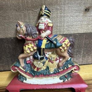 May include: A painted cast iron nutcracker figurine depicting a soldier in a black hat and red uniform riding a brown horse. The base features a small teddy bear, a drum, and green holly leaves, all mounted on a solid red rectangular metal stand.