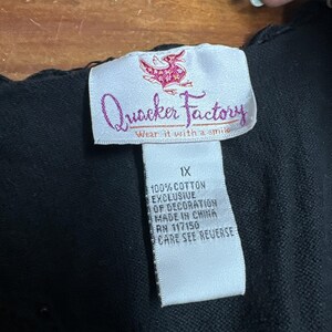 May include: A white clothing tag on a black garment featuring a pink duck logo and the text Quacker Factory Wear it with a smile. The tag lists the size as 1X, 100% cotton, exclusive of decoration, made in China, and RN 117150.