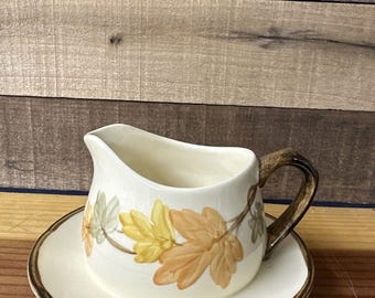 May include: A white ceramic gravy boat with a brown handle and a matching saucer. The boat is decorated with a pattern of orange, yellow, and green leaves.