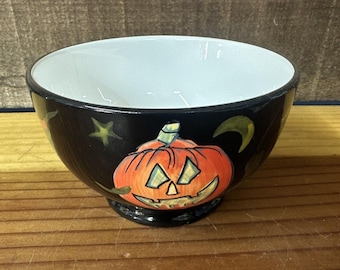 May include: Black ceramic bowl with a white interior. The exterior features a painted orange jack-o'-lantern, green stars, and a crescent moon. A festive Halloween-themed bowl.