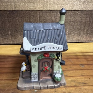 May include: A ceramic miniature Spring House building featuring a dark gray roof with white snow accents, a light green stone-textured facade, a brown arched door with a red bow wreath, a small white duck, and a black lantern hanging by the door.