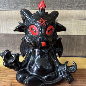 May include: Black ceramic figurine of a demon with red eyes, horns, and a pentagram on its forehead. The demon is sitting in a meditative pose with its hands in a mudra.