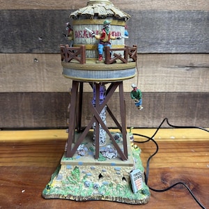 May include: A miniature water tower figurine featuring a Deadwood sign, skeletal figures in western attire, and a hanging figure. The structure has a stone base, wooden support beams, and a sign that reads Tower Continues Keep Out.